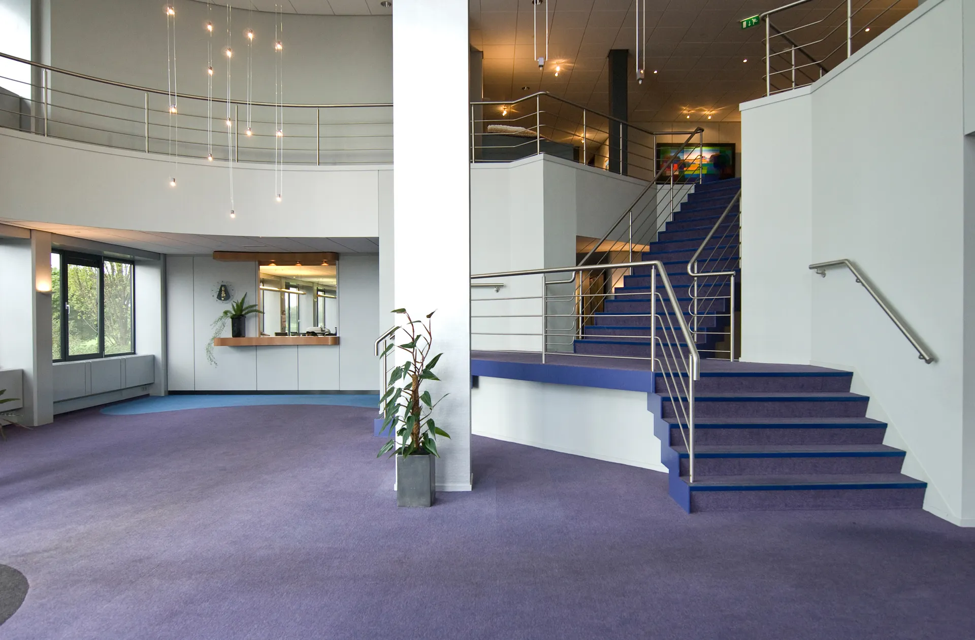 Een ruime, moderne kantoorlobby met een grote open ruimte, witte muren, blauwe vloerbedekking, een trap met leuning en een plant in een pot.