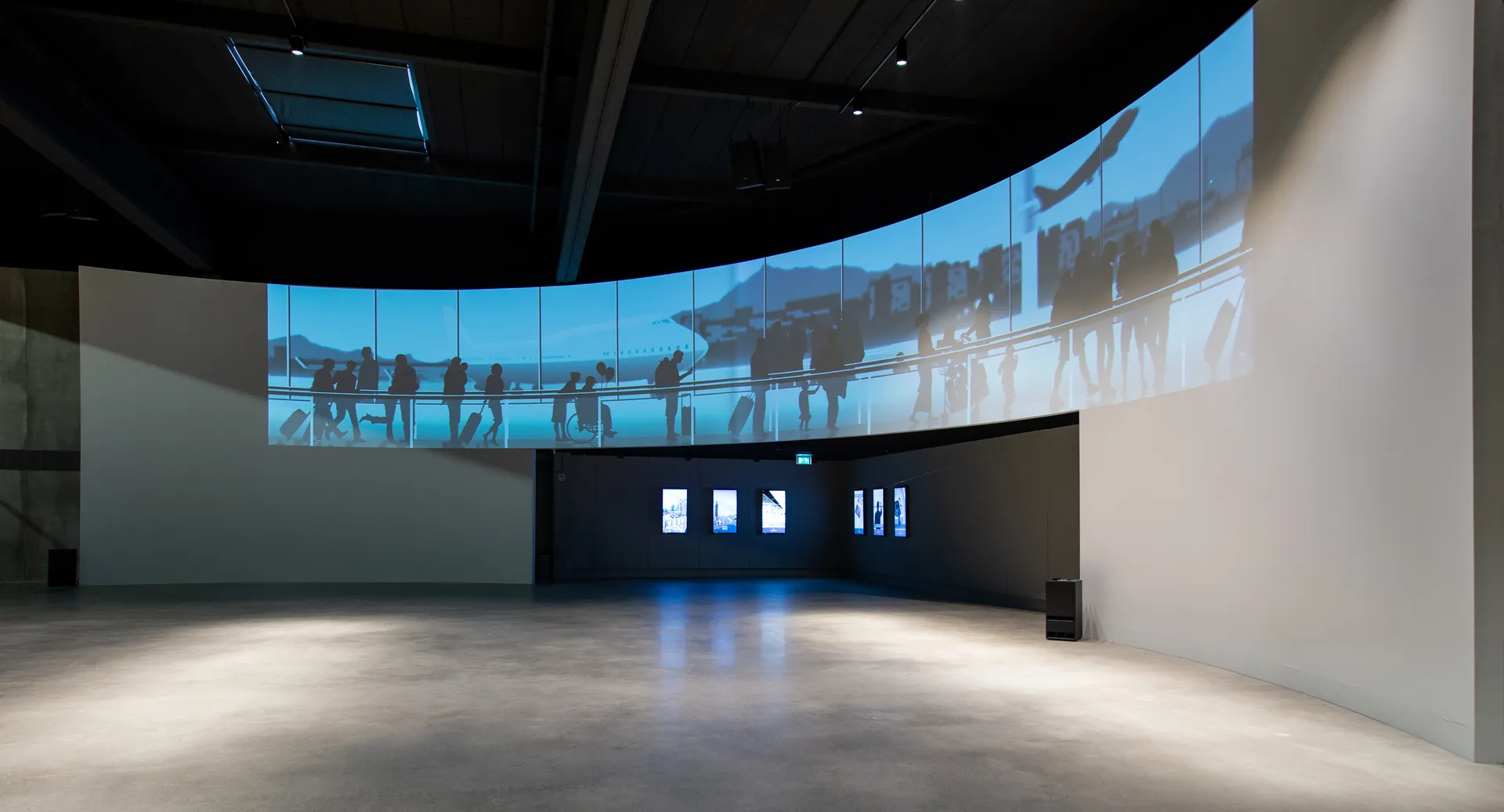 Een grote, open ruimte met een gebogen scherm waarop een video van mensen op een brug wordt getoond, omgeven door witte muren en een betonvloer.