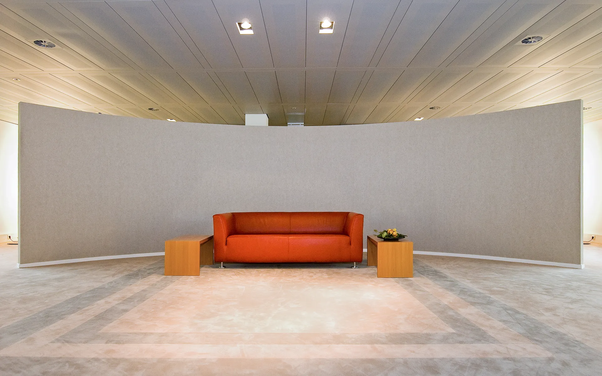 Een moderne wachtruimte in een kantoor met een comfortabele oranje bank, een houten tafeltje en een grote witte muur, voor een professionele uitstraling.