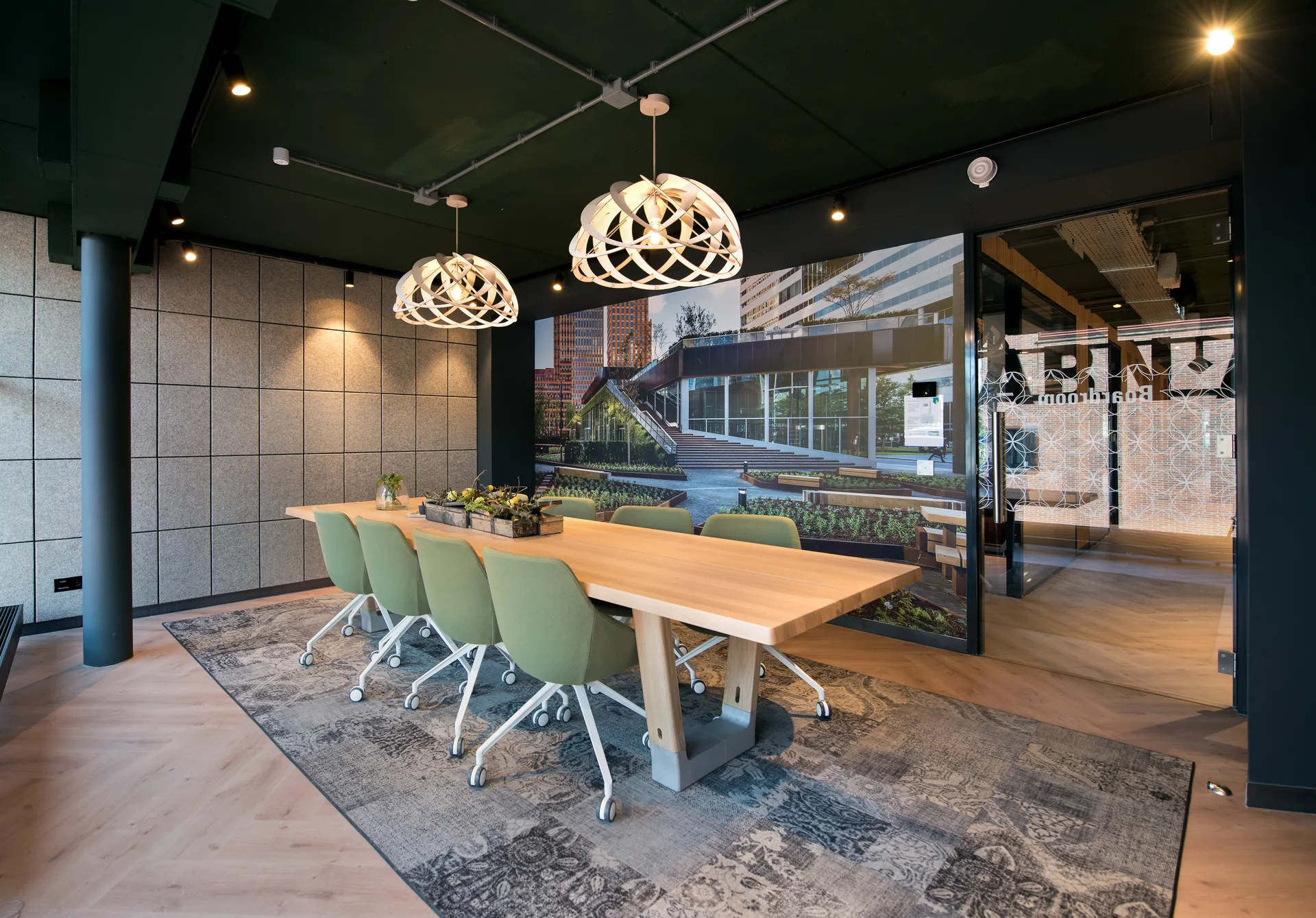 Een moderne vergaderruimte met een grote houten tafel, groene stoelen en een groot scherm aan de muur.