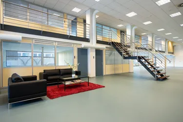 Een moderne kantoorruimte met een zwarte bank, rood vloerkleed en glazen salontafel, omgeven door grote ramen en een trap.