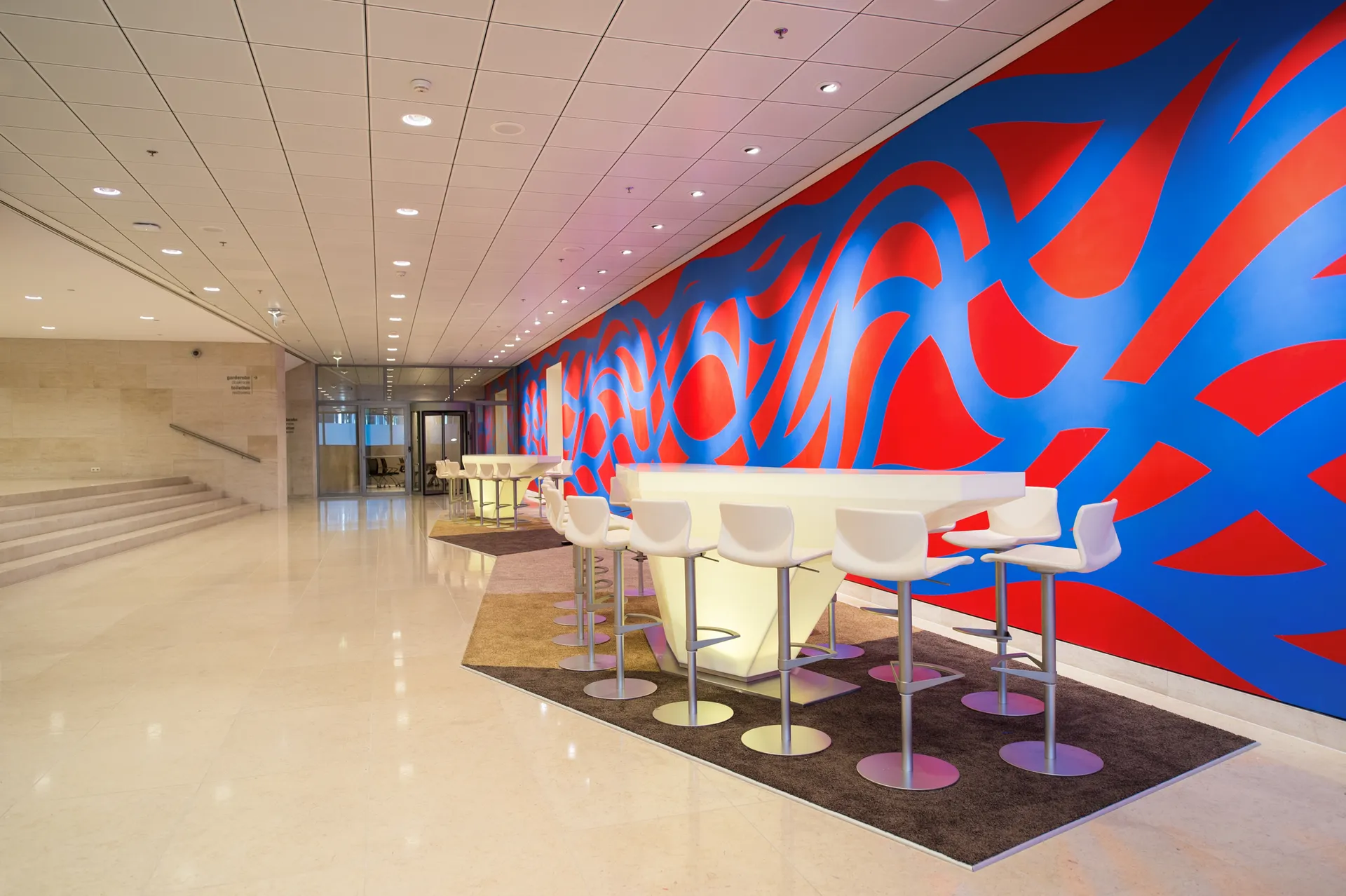 Een moderne wachtruimte vol kleur met een abstracte rood-wit-blauwe muurschildering en speelse wandtafels om aan te zitten.